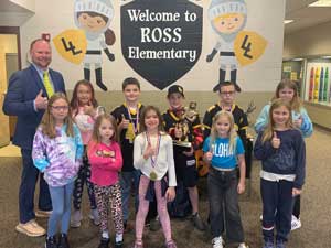 An adult with students. Some are wearing medals and one is holding a trophy. They are under a wall painting that reads Welcome to Ross Elementary